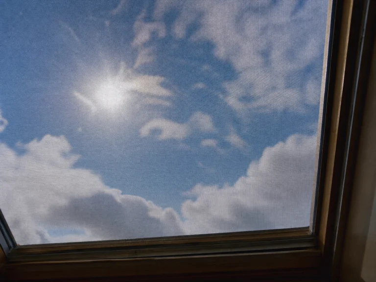 How to Clean Skylights Safely (Without Streaks or Scratches) 2 Sunlight shines through a screened skylight onto a blue sky with soft white clouds, framed by dark wood; a peaceful, airy scene.
